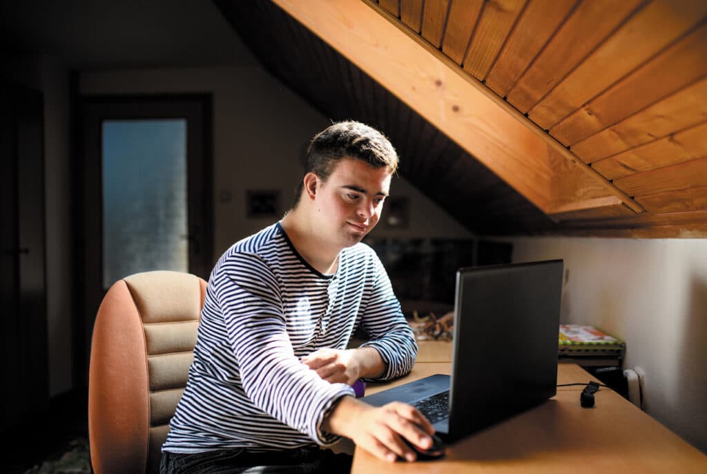 Young man with Down syndrome sitting at a desk in a cozy, attic room, using a laptop, illuminated by natural light.