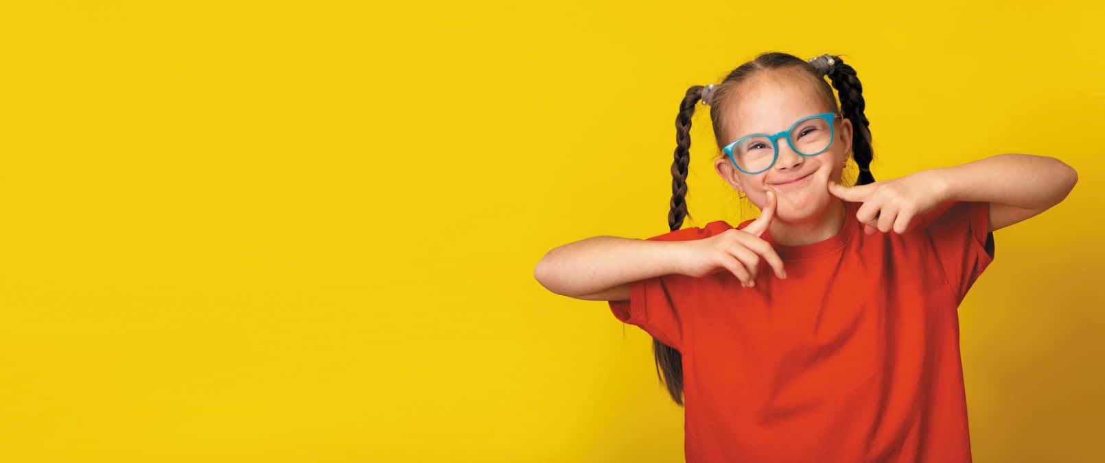 Smiling young girl with Down syndrome wearing blue glasses and a red shirt, making a playful gesture against a bright yellow background.