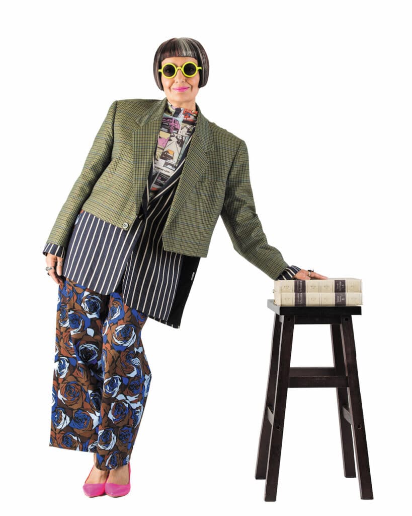 Visionary eyewear women leader Sue Randhawa in a stylish outfit, including thick yellow-framed sunglasses, leaning against a pair of books on a stool, against a white background.