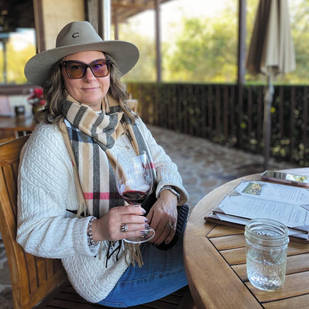 Christine Zeggil sitting on a restaurant patio, with a glass of water and a menu in front of her. She is holding a glass with a red beverage.